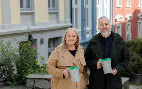 En blond kvinne i beige kåpe og en mann med skjegg og svart frakk står med bøsser utenfor hus i en by. 
