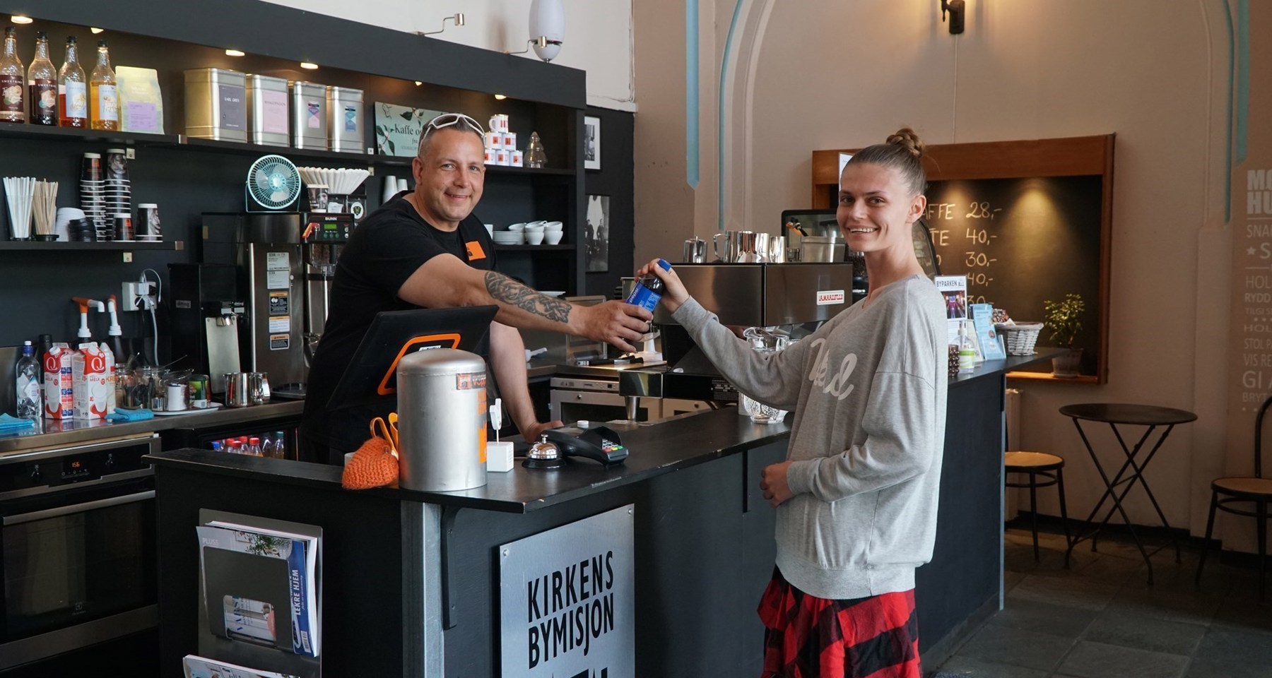 Kafé på Kirkens Bymisjon, en mann står bak disken og serverer en brus til en kvinne på andre siden.