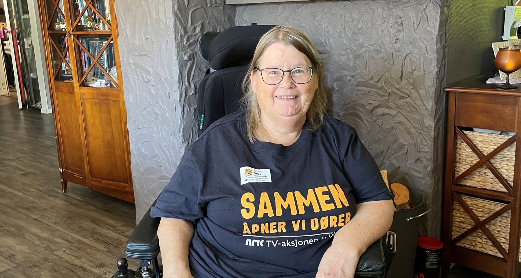 Kvinne sitter i rullestol inne i en stue. Hun har på seg TV-aksjons-trøye med skriften Sammen åpner vi dører. 