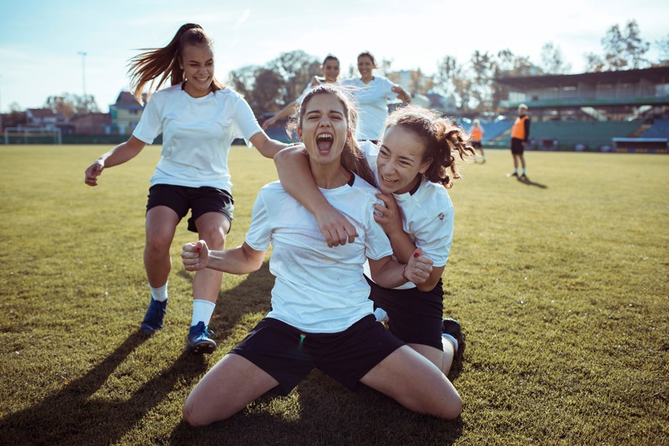To jenter sitter på bakken med uttryk at de feirer fotballmål. En annen jente kommer løpende bak. Alle har like, hvite fotballdrakter.