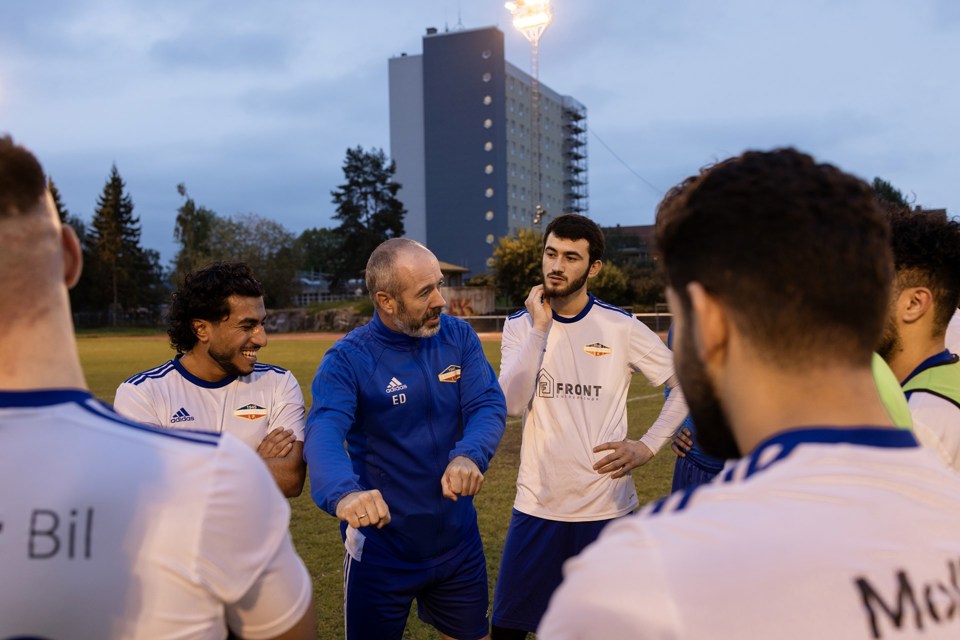 En gjeng menn samlet på en fotballbane. En trener i blå drakt snakker, mens spillere i hvite drakter lytter.