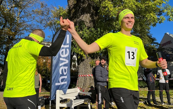 To menn i neongule tskjorter holder hverandre i hendene hevet, en med stafettpinne i hånden 