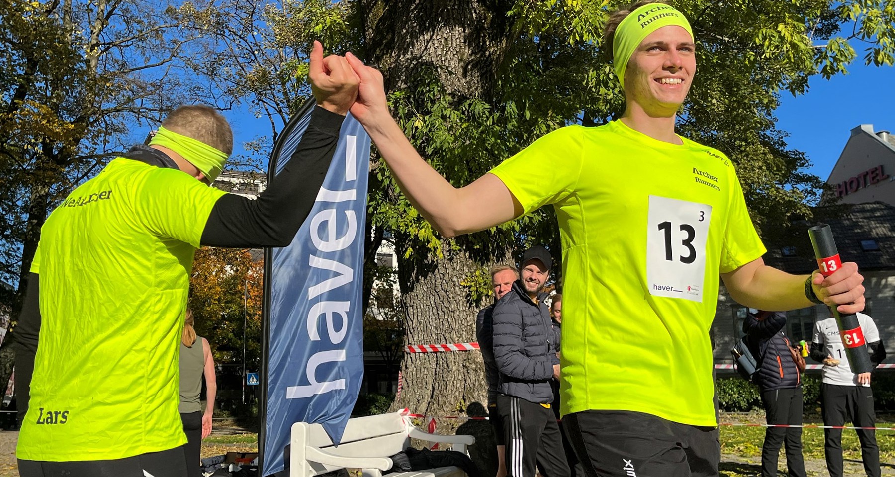 To menn i neongule tskjorter holder hverandre i hendene hevet, en med stafettpinne i hånden 