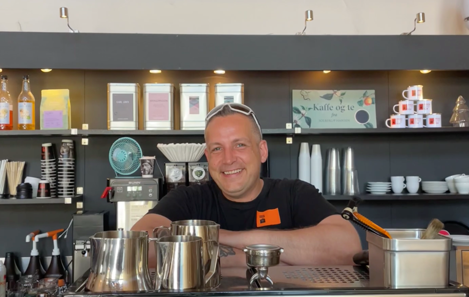 En mann som smiler og står bak disken på en kafé.
